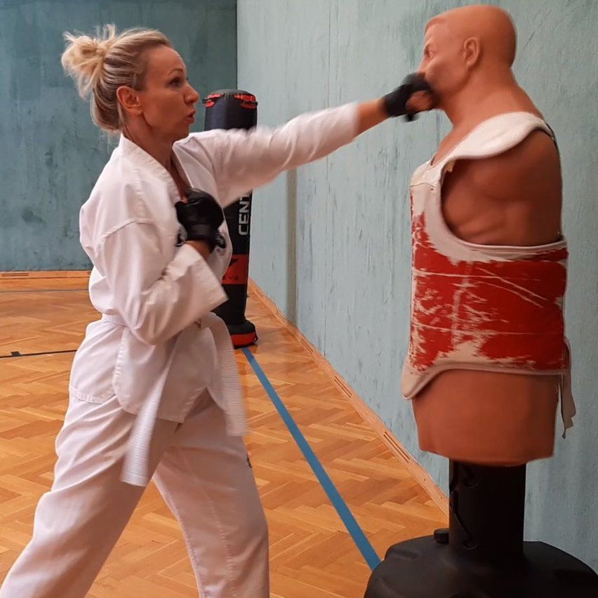 A woman dressed in a white karate uniform practices punches on a mannequin in a gym.