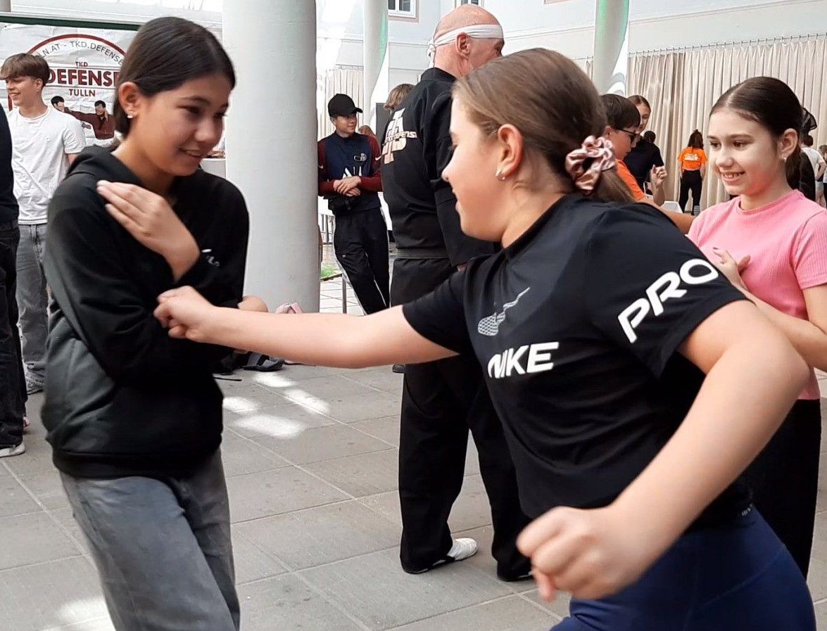 Zwei junge Frauen in schwarzen Nike-Shirts lächeln und tanzen zusammen in einem Innenraum. Hinter ihnen beobachtet ein Mann mit Kopfband.