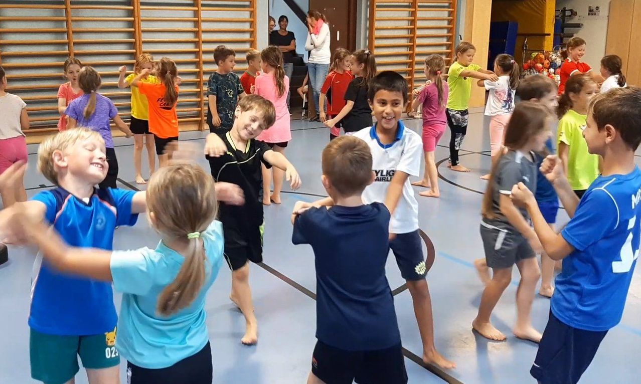 Eine Gruppe von Kindern in einer Turnhalle, einige tanzen, während andere zuschauen. Die Kinder tragen verschiedene farbige Shirts und Shorts.