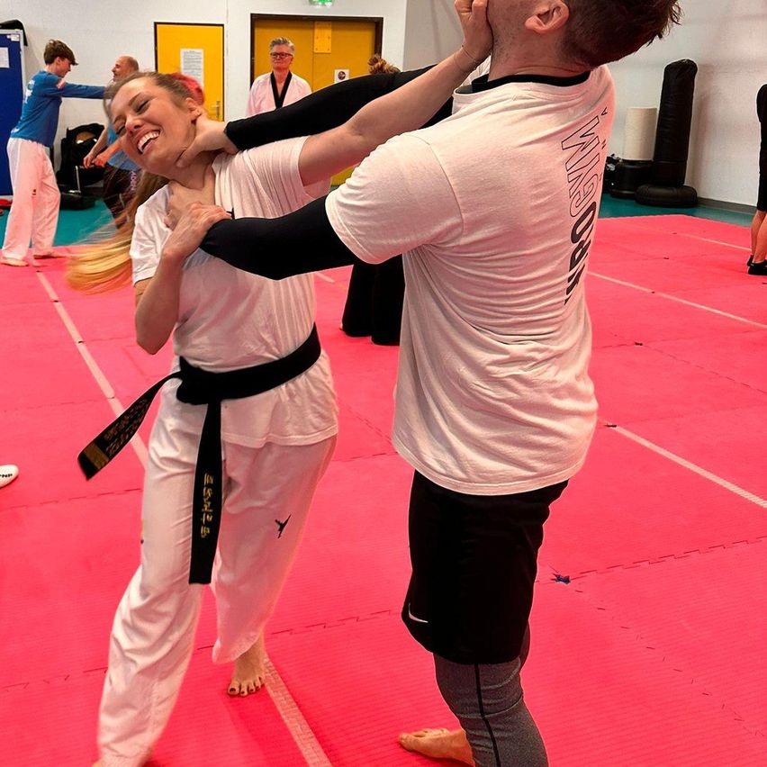 Eine Frau in einem weißen Karate-Anzug mit einem schwarzen Gürtel übt Kampfsport mit einem Mann in einem weißen T-Shirt und schwarzen Shorts auf einem roten Teppich.