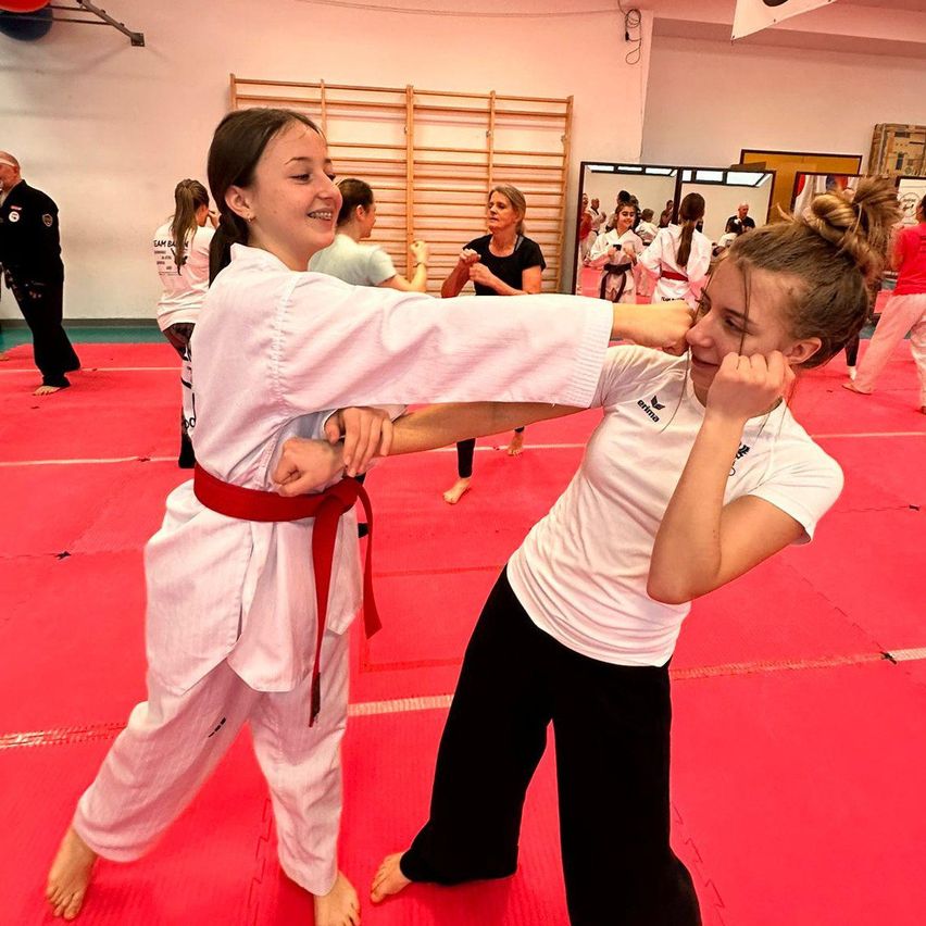 Zwei junge Frauen in weißen Kampfsportuniformen üben eine Grappling-Technik auf einem roten Matten. Eine Frau hält den Arm der anderen, während die andere sich auf einen Schlag vorbereitet. Andere Schüler und Ausbilder sind im Hintergrund.