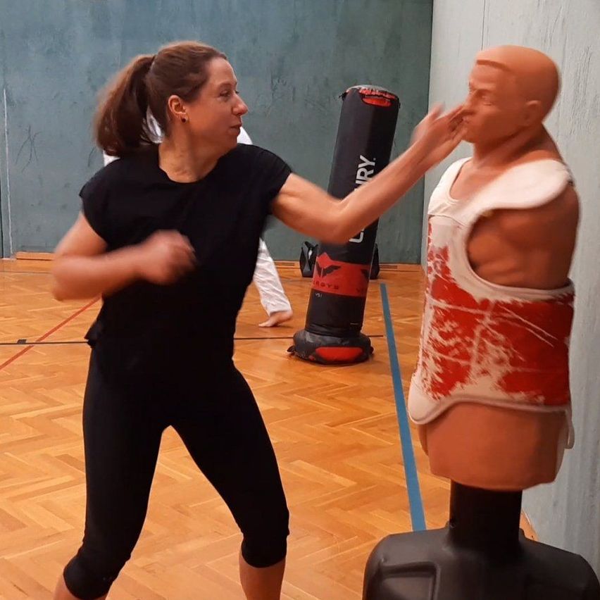 Eine Frau in schwarzer Trainingskleidung übt Kampfsport, indem sie eine Puppe in einem Fitnessstudio schlägt. Ein Boxsack und eine weitere Person sind im Hintergrund zu sehen.