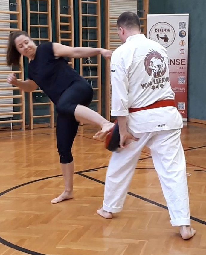 Eine Frau in schwarzer Sportbekleidung führt einen hohen Tritt aus, während ein Mann in einem weißen Kampfsportanzug einen roten Beutel in einer Turnhalle hält.