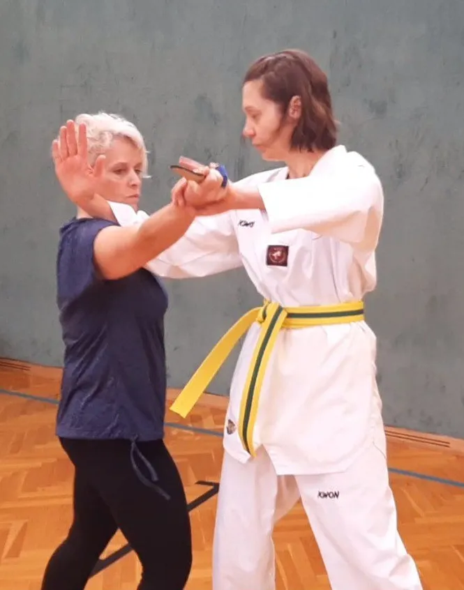 Zwei Frauen in einem Fitnessstudio, eine in einem blauen Shirt und schwarzen Hosen, die andere in einem weißen Karate-Anzug mit einem gelben Gürtel, üben Kampfsport.