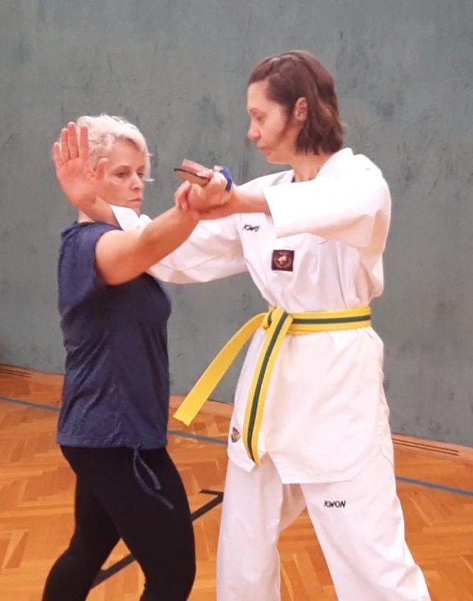 Zwei Frauen in einem Fitnessstudio, eine in einem blauen Shirt und schwarzen Hosen, die andere in einem weißen Karate-Anzug mit einem gelben Gürtel, üben Kampfsport.