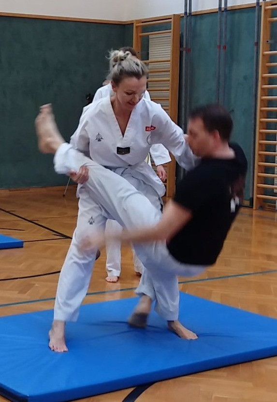 Eine Frau in einer weißen Karate-Uniform führt einen hohen Tritt auf einen Mann in einem schwarzen Shirt auf einem blauen Matten in einem Fitnessstudio durch.