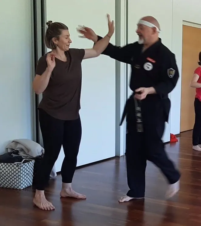Eine Frau in einem braunen T-Shirt und schwarzen Leggings übt Kampfkunst mit einem Mann in einem schwarzen Gi und Stirnband. Sie sind barfuß in einem Studio mit Holzböden.