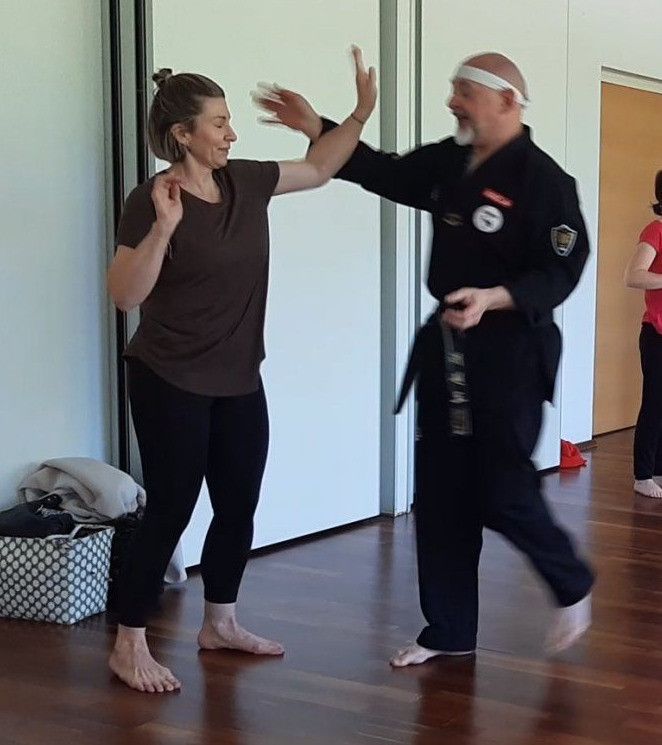 Eine Frau in einem braunen T-Shirt und schwarzen Leggings übt Kampfkunst mit einem Mann in einem schwarzen Gi und Stirnband. Sie sind barfuß in einem Studio mit Holzböden.