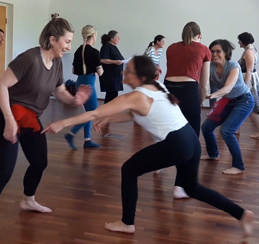 Eine Gruppe von Frauen übt in einem Studio Tanz, wobei eine Frau die anderen in einer verspielten Interaktion anführt.
