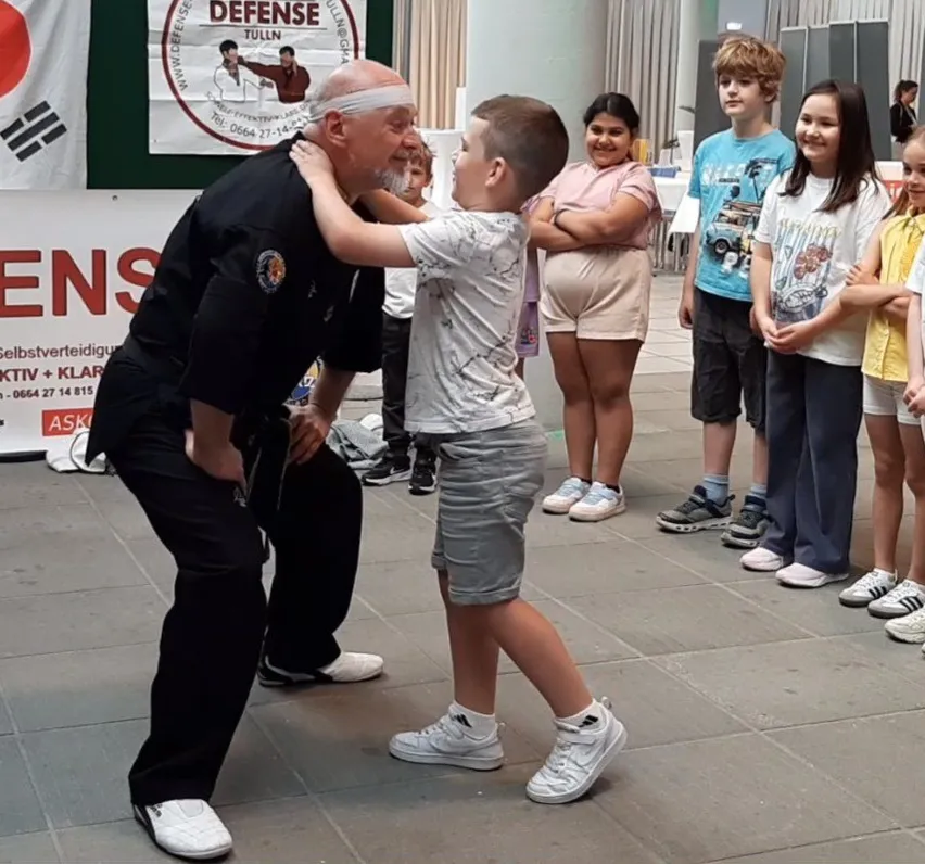 Ein älterer Mann unterrichtet einen jungen Jungen in Kampfkunst in einem Raum mit einem Banner im Hintergrund.