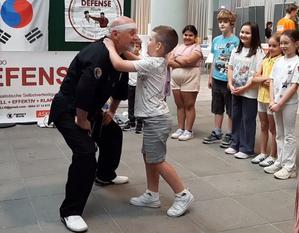 Ein älterer Mann unterrichtet einen jungen Jungen in Kampfkunst in einem Raum mit einem Banner im Hintergrund.