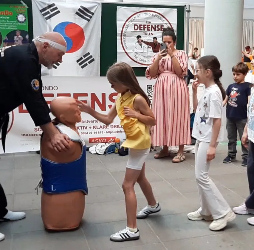 Ein Mann in einem schwarzen Kampfsportanzug unterrichtet ein junges Mädchen, wie man einen Dummy schlägt. Sie sind von anderen Kindern und Erwachsenen umgeben.