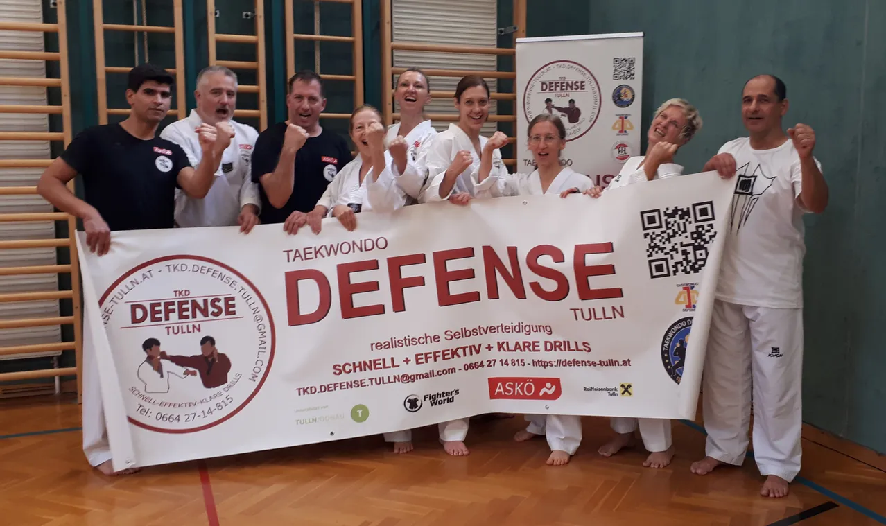 Eine Gruppe von Menschen in weißen Kampfsportuniformen posiert für ein Foto. Sie halten ein Banner mit der Aufschrift 'Verteidigung' in großen roten Buchstaben.