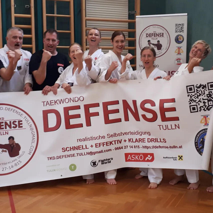 Eine Gruppe von Menschen in weißen Kampfsportuniformen posiert für ein Foto. Sie halten ein Banner mit der Aufschrift 'Verteidigung' in großen roten Buchstaben.
