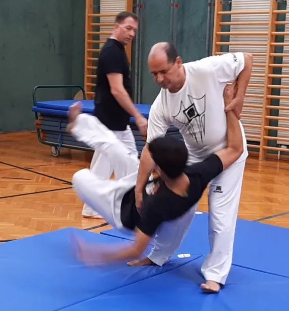 Ein Mann in einem weißen Outfit unterrichtet einen jungen Mann in einem schwarzen Outfit, wie man eine Kampfkunstbewegung auf einem blauen Teppich in einem Fitnessstudio durchführt.