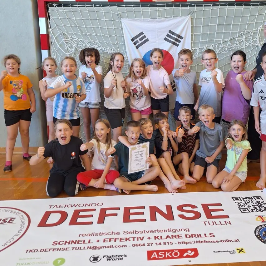 Eine Gruppe von Kindern und zwei Erwachsenen posiert für ein Foto in einer Turnhalle. Sie alle lächeln und einige halten Urkunden. Ein Banner mit dem Wort 'Verteidigung' liegt auf dem Boden.