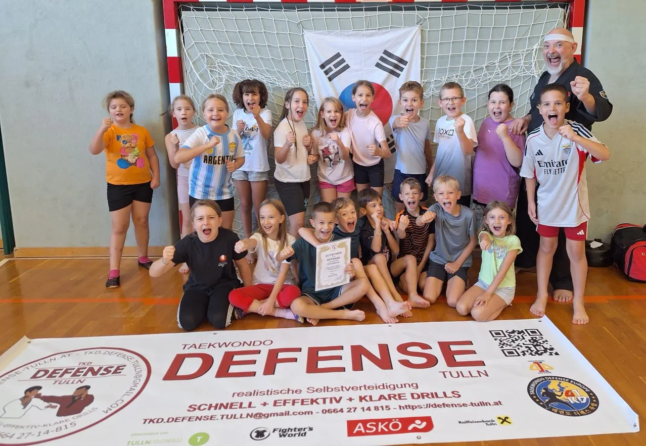 Eine Gruppe von Kindern und zwei Erwachsenen posiert für ein Foto in einer Turnhalle. Sie alle lächeln und einige halten Urkunden. Ein Banner mit dem Wort 'Verteidigung' liegt auf dem Boden.