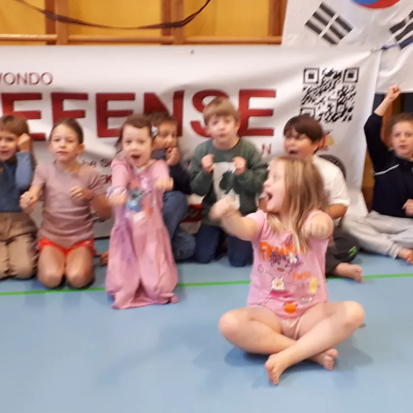 Eine Gruppe von Kindern sitzt auf dem Boden in einer Turnhalle, einige tragen rosa Kleider, und ein Banner mit den Worten 'Taekwondo Defense' hängt an der Wand hinter ihnen.