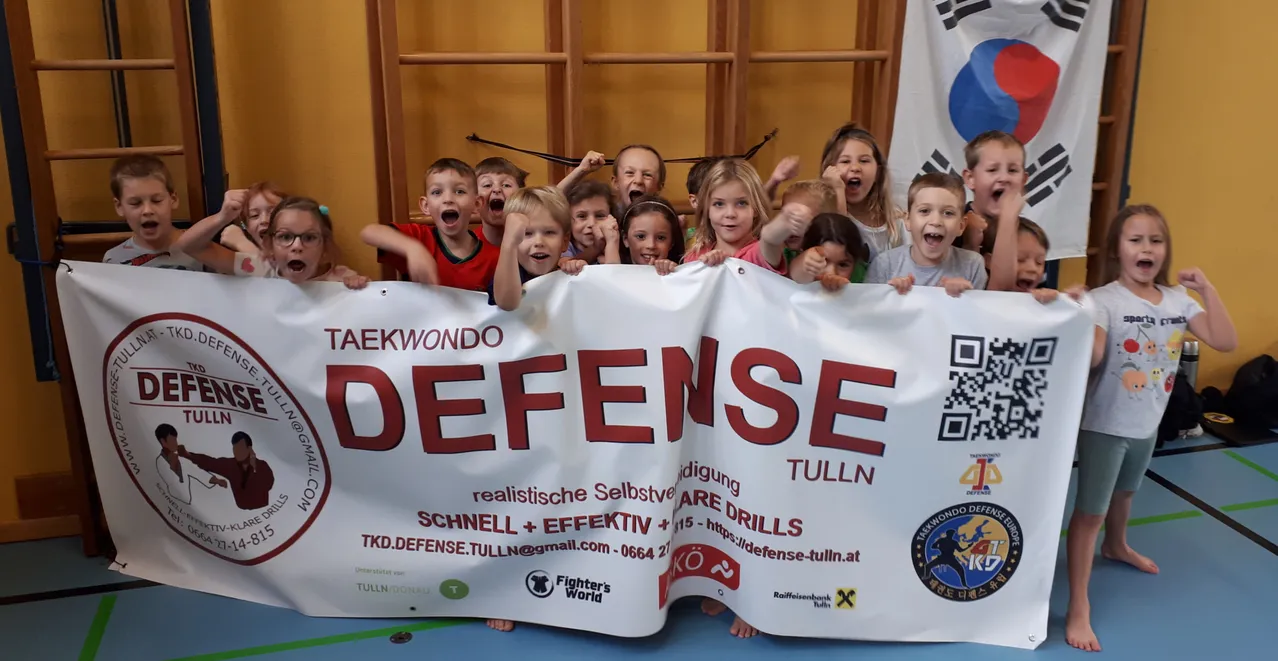 Eine Gruppe von Kindern in einem Kampfsportstudio, die ein Banner halten, das 'Taekwondo Defense Tulln' sagt. Sie tragen alle Uniformen und scheinen Kampfsport zu üben.