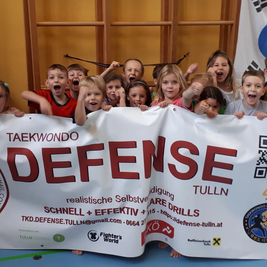Eine Gruppe von Kindern in einem Kampfsportstudio, die ein Banner halten, das 'Taekwondo Defense Tulln' sagt. Sie tragen alle Uniformen und scheinen Kampfsport zu üben.