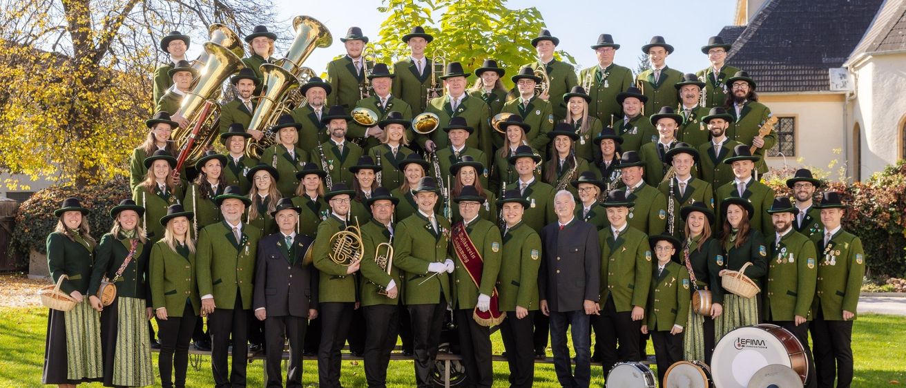 Eine große Gruppe von Musikern in grünen Uniformen posiert mit verschiedenen Instrumenten, darunter Posaunen, Trompeten und Trommeln, auf einem Rasenfeld.