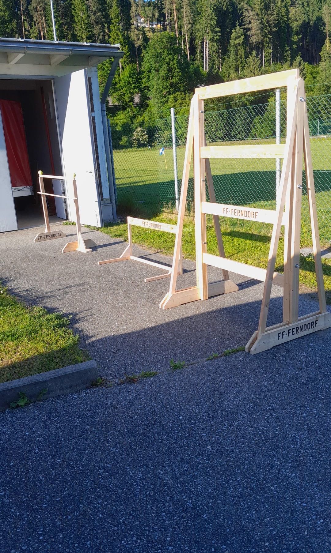 Drei Holzhurdele mit der Aufschrift FF-Ferndorf stehen auf einer Betonfläche mit einem Rasenbereich und einem Zaun im Hintergrund.