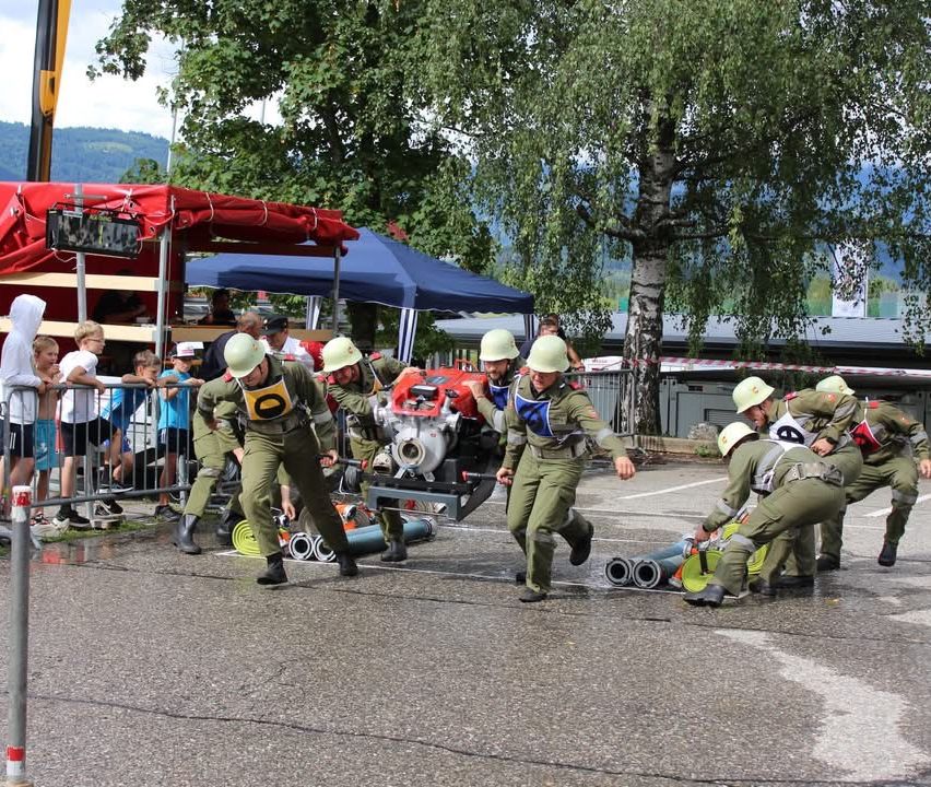 Eine Gruppe von Menschen in Uniformen und Helmen rennt und schiebt ein Fahrzeug mit einer Maschine darauf. In der Nähe befinden sich Zelte und eine Menge zuschauender Menschen.
