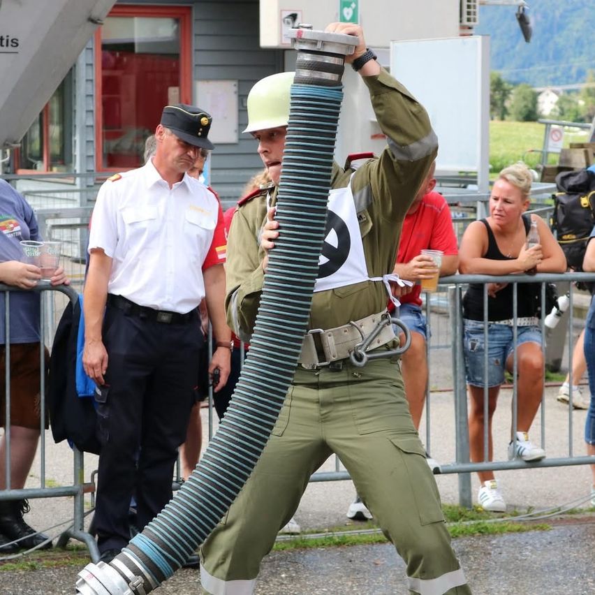 Ein Feuerwehrmann in Uniform hält einen großen Schlauch in der Hand und steht vor einer Menschenmenge. Die Menge steht hinter einem Metallzaun. Im Hintergrund befindet sich ein Gebäude mit einem roten Fenster und Bergen.