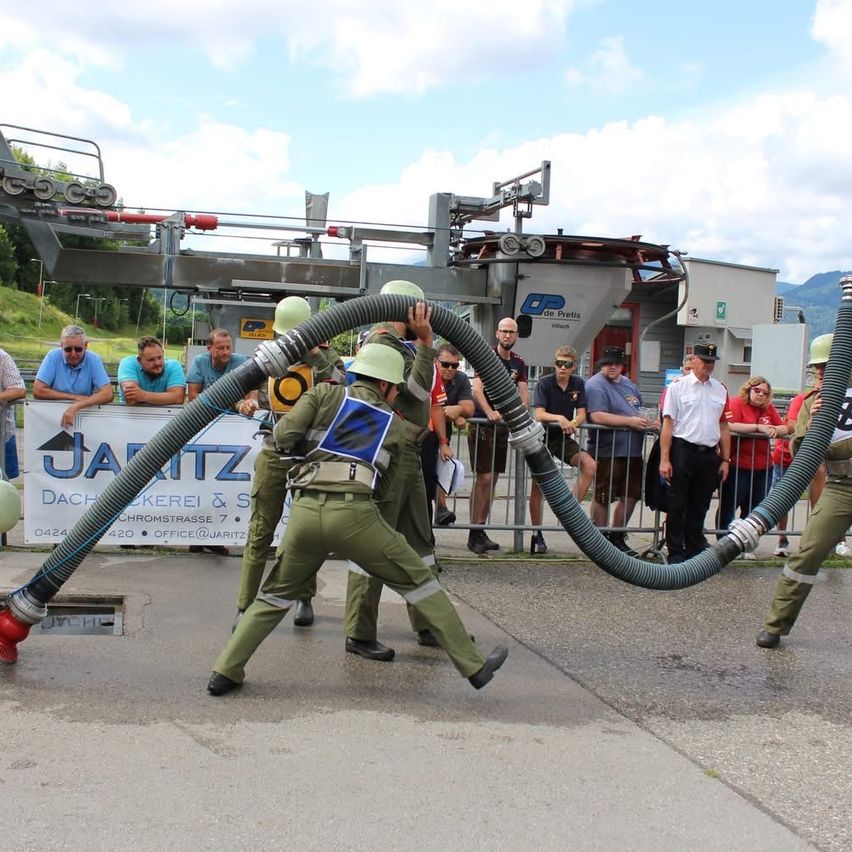 Feuerwehrleute demonstrieren eine Wasserkanone. Sie tragen Helme und Uniformen. Ein Publikum beobachtet sie.
