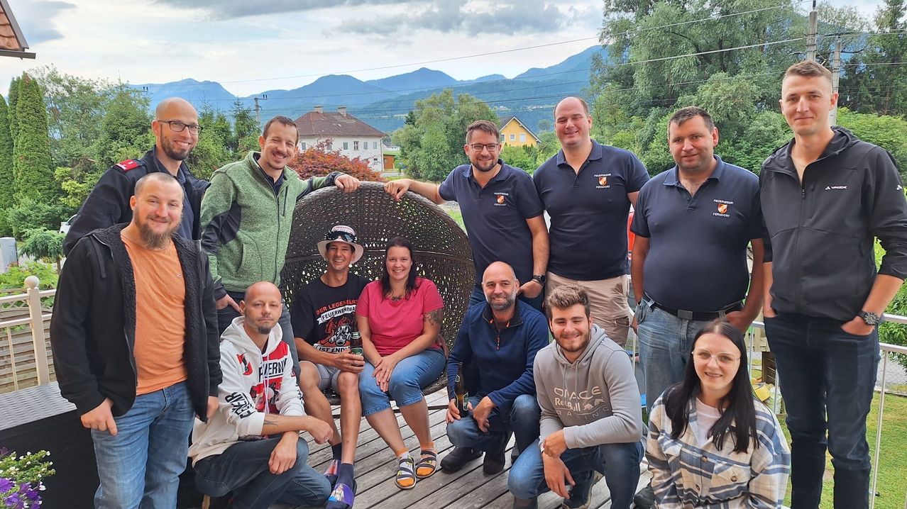 Eine Gruppe von Menschen, einige in Jacken und andere in Freizeitkleidung, posiert für ein Foto auf einer Holzterrasse mit Bergen im Hintergrund.
