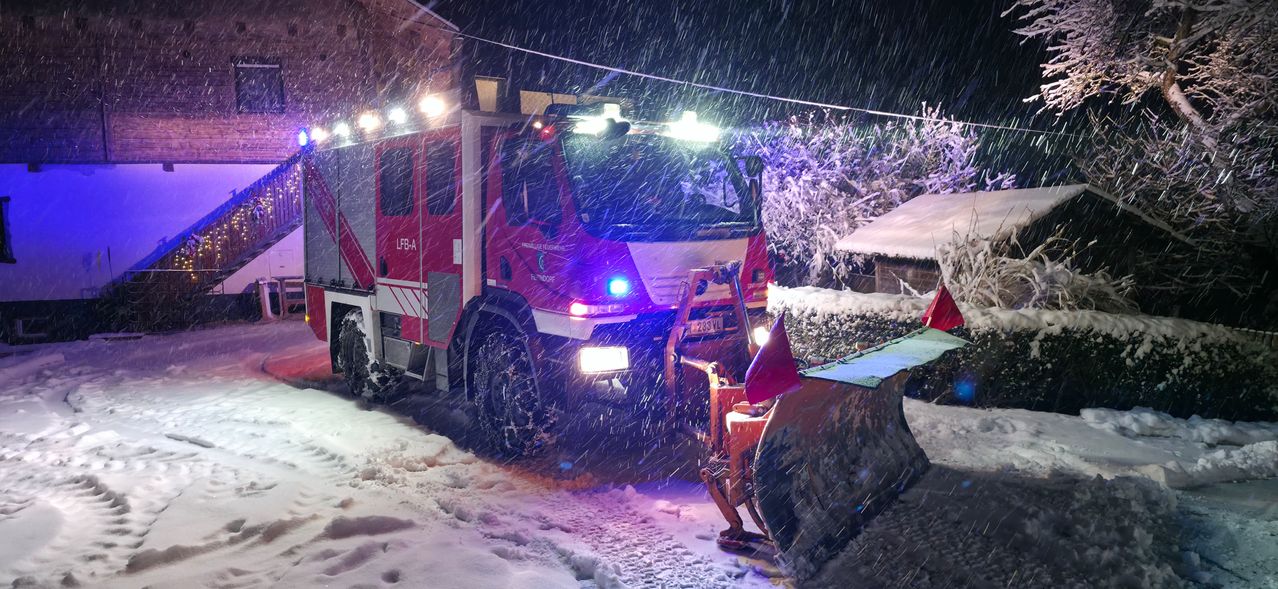 Ein roter Feuerwehrwagen mit einem Schneepflug räumt in starkem Schneefall eine Straße vom Schnee frei.
