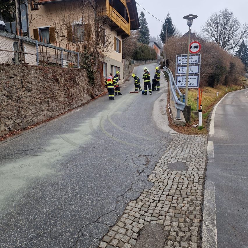 Eine Gruppe von Feuerwehrleuten in Schutzausrüstung reinigt die Straße mit Besen auf einer kleinen Straße. Ein Schild ist an einem Pfosten auf der rechten Seite montiert.