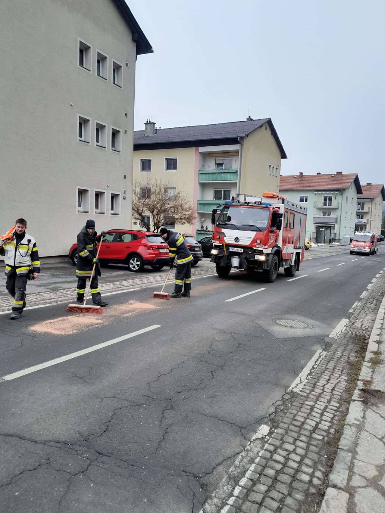 Drei Feuerwehrleute fegen die Straße mit einem Besen. Ein roter Feuerwehrwagen ist dahinter geparkt. Auf der gegenüberliegenden Straßenseite befinden sich zwei Gebäude, eines mit einem roten Dach. Ein rotes Auto ist vor dem Gebäude geparkt.