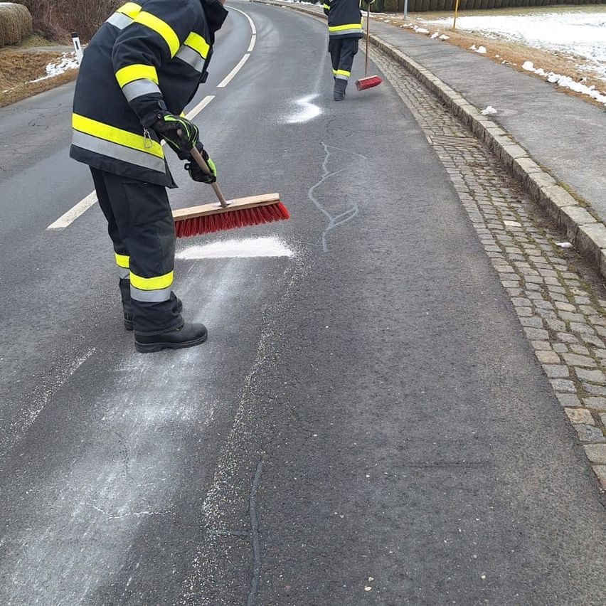 Zwei Personen, wahrscheinlich Feuerwehrleute, sind auf der Straße und reinigen sie mit Besen. Sie tragen Schutzkleidung. Die Straße hat weiße Markierungen.