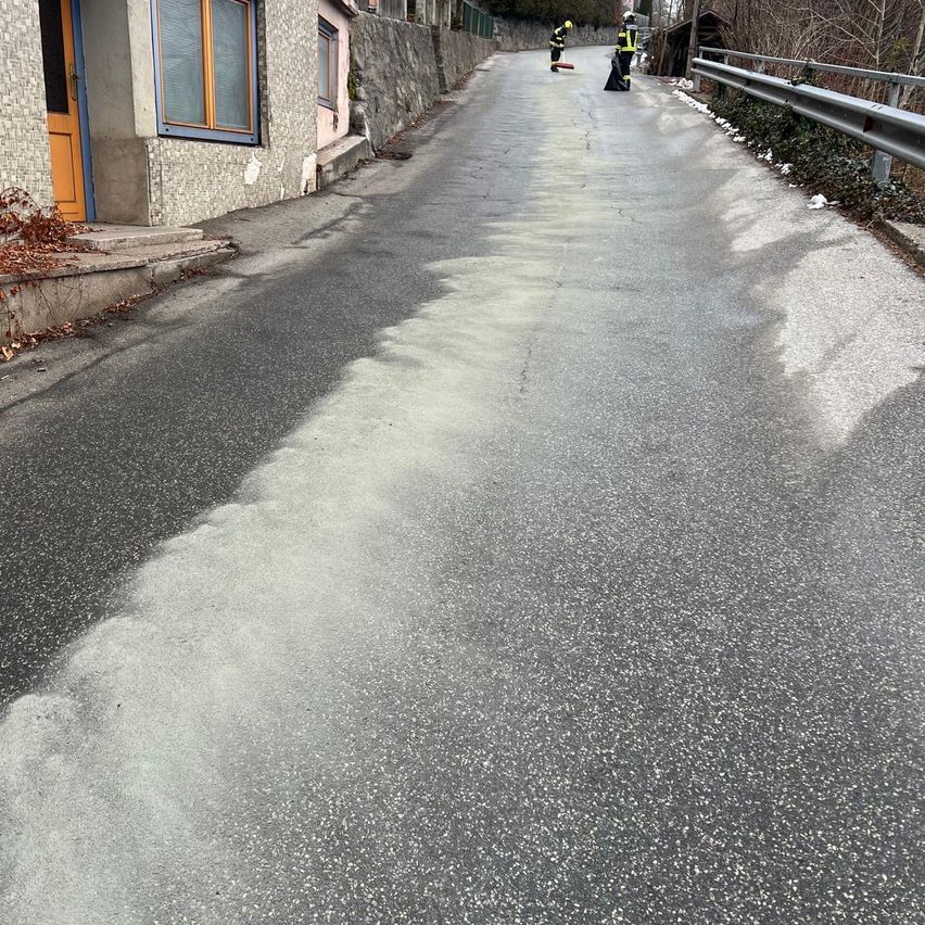 Zwei Arbeiter sind auf einer Straße mit einem Metallgeländer auf der Seite, und sie verteilen weißes Pulver. Es gibt Gebäude mit Glasfenstern auf der linken Seite.