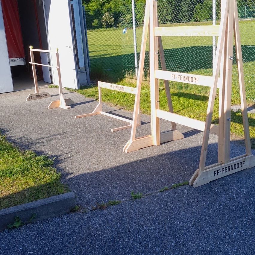 Drei Holzhürdchen mit der Aufschrift FF-Ferndorf sind auf einem Bürgersteig in der Nähe eines Gebäudes mit offener Tür aufgestellt. Hinter den Hürdchen befindet sich ein Rasenfeld mit Zaun.