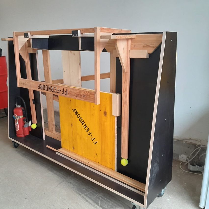 Eine hölzerne Struktur mit einer gelben Tür, beschriftet mit "FENCER", steht auf Rädern. Es gibt einen Feuerlöscher und Tennisbälle in der Nähe. Die Struktur befindet sich in einem Raum mit weißen Wänden.