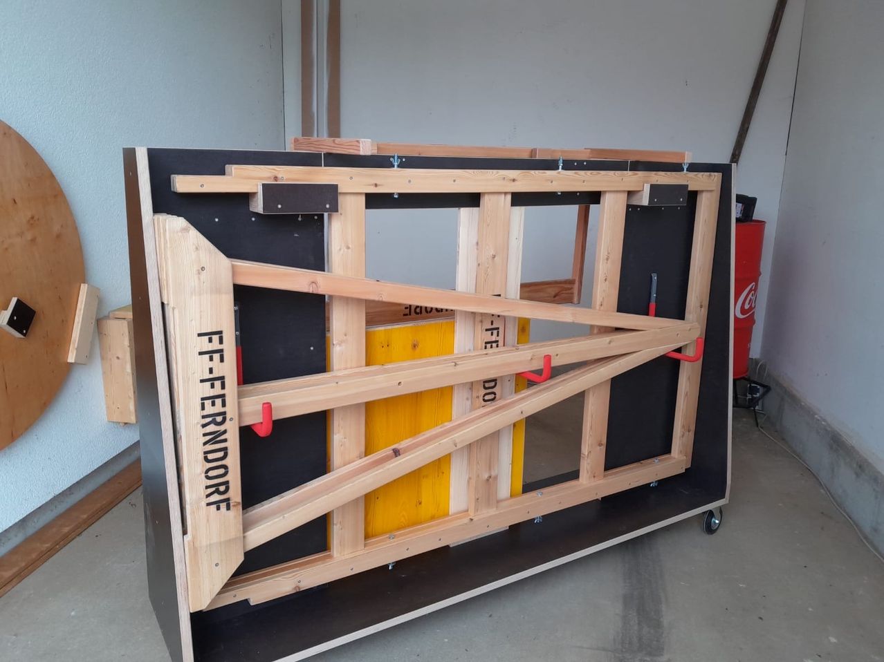 Eine Holzstruktur mit dem Text FF-FERNDORF darauf, befestigt an einem schwarzen Sockel, mit roten Griffen. Dahinter steht eine gelbe Struktur mit dem Text FE-TE DO. Rechts davon steht ein roter Coca-Cola-Fass. Links davon ist eine weiße Wand.