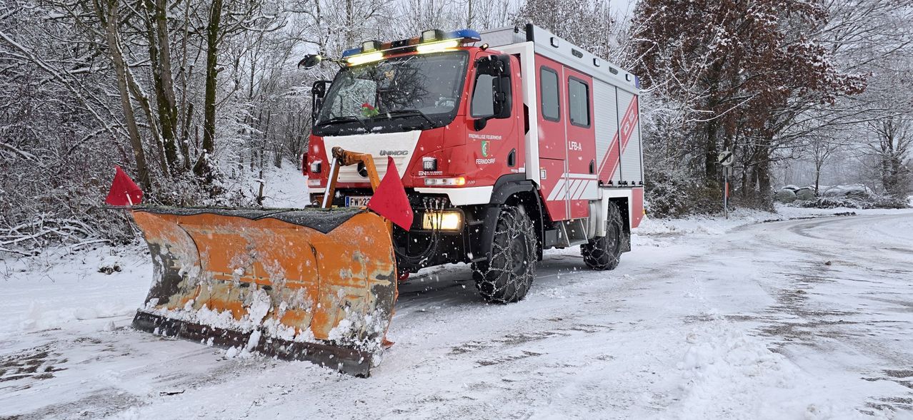 Ein roter Feuerwehrwagen mit einem Schneepflug fährt auf einer verschneiten Straße. Der Wagen hat ein Nummernschild und das Wort UNIMOG ist auf der Vorderseite.