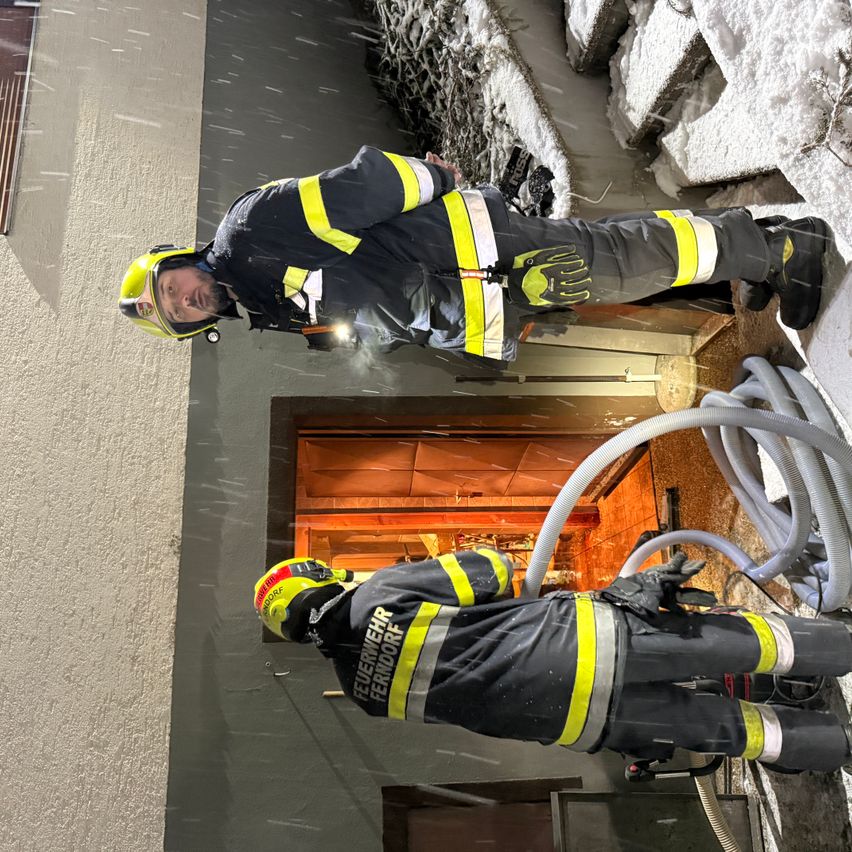 Zwei Feuerwehrleute klettern an einer Wand. Einer steht und hält einen Schlauch, während der andere unter ihm ist. Sie tragen beide Helme und Uniformen.