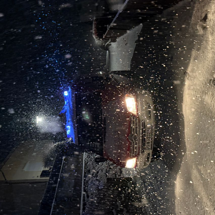 Ein Auto mit blauen Lichtern auf der Front fährt bei Nacht auf einer verschneiten Straße. Die Scheinwerfer sind eingeschaltet, und das Auto ist mit Schnee bedeckt.