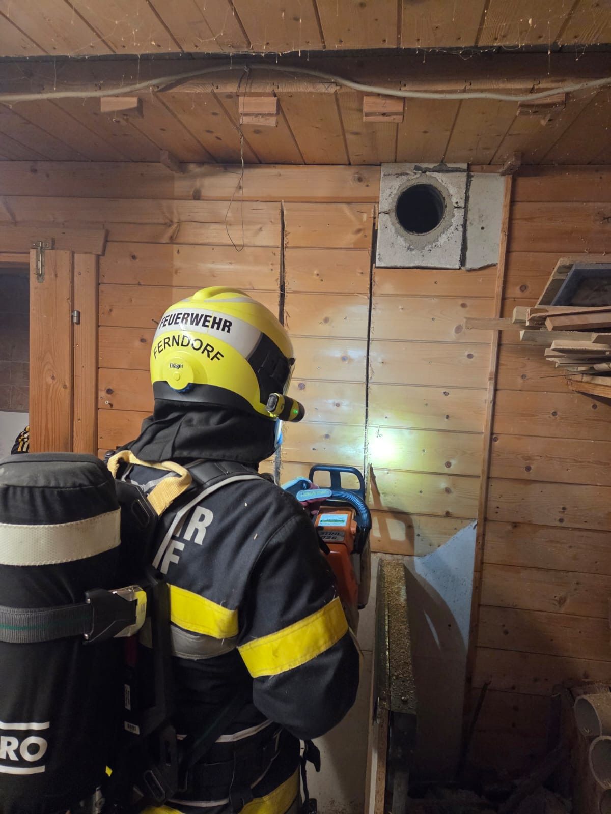 Ein Feuerwehrmann in voller Ausrüstung steht in einem Holzgebäude und säg mit einer Kettensäge durch ein Loch in der Wand.