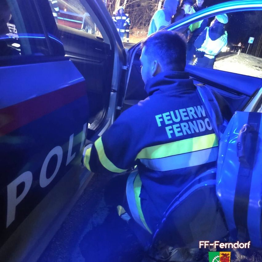 Ein Feuerwehrmann hilft einer Person in einem Polizeiwagen. Die Tür des Wagens ist geöffnet, und auf der Seite des Polizeiwagens steht 'POLIZEI'.
