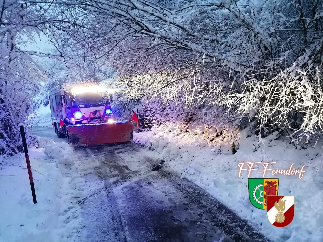 Ein Schneepflug mit blauen Lichtern räumt eine verschneite Straße. Schneebedeckte Bäume säumen die Seiten der Straße.