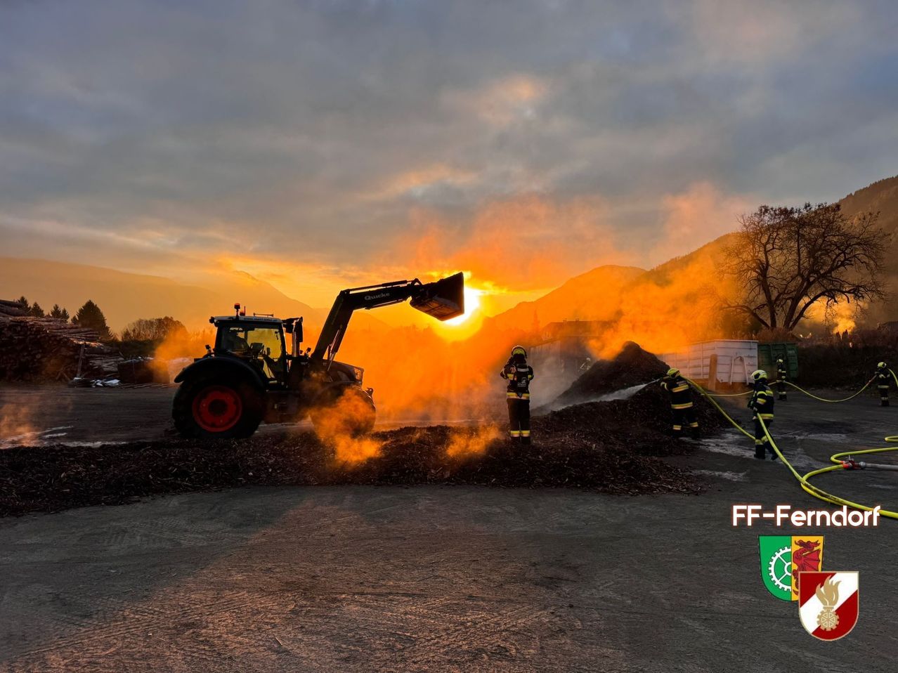 Firefighters are battling a blaze near a tractor in a rural setting at sunset. Smoke billows from the tractor and the pile of wood being extinguished.