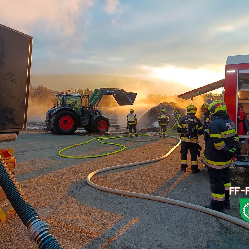 Firefighters are standing by a truck and hose, with a tractor in the background and a setting sun.