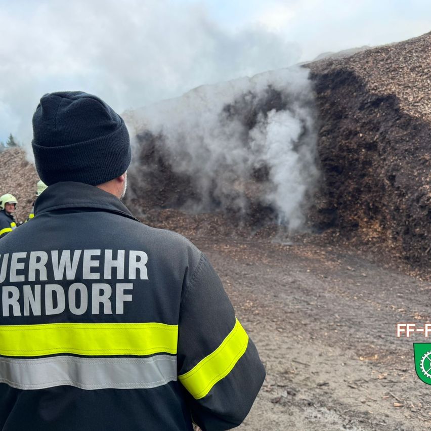 A firefighter with a beanie looks towards a pile of smoldering wood. A plume of smoke rises from the pile.