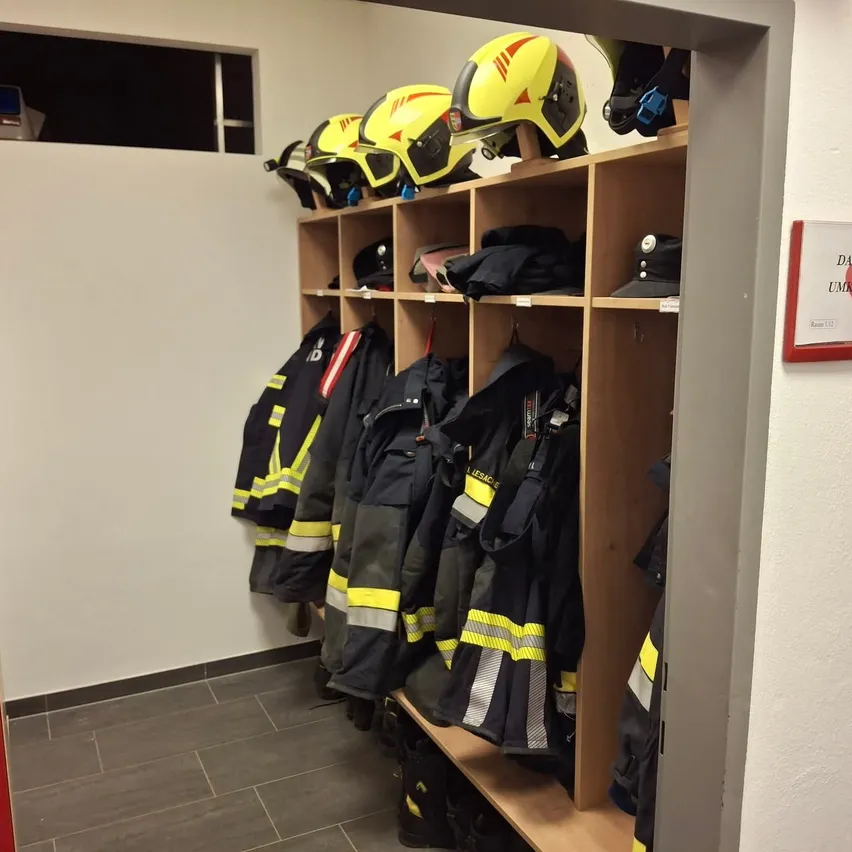 Ein Raum mit einem Holzregal, das Helme und Feuerwehruniformen enthält, mit einer weißen Wand und einem gefliesten Boden.