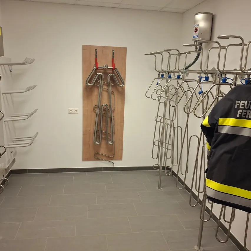 Ein leerer Umkleideraum mit einem Feuerwehrmannhemd an einem Haken. Ein hölzerne Halterung mit gebogenen Eisenfiguren ist an der Wand montiert.