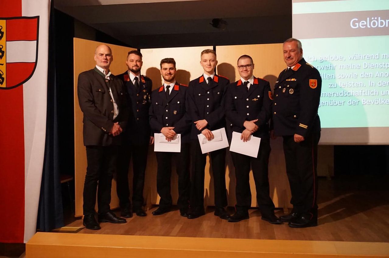 Five men in uniforms stand on a stage holding certificates. They are smiling and appear to be celebrating an achievement.